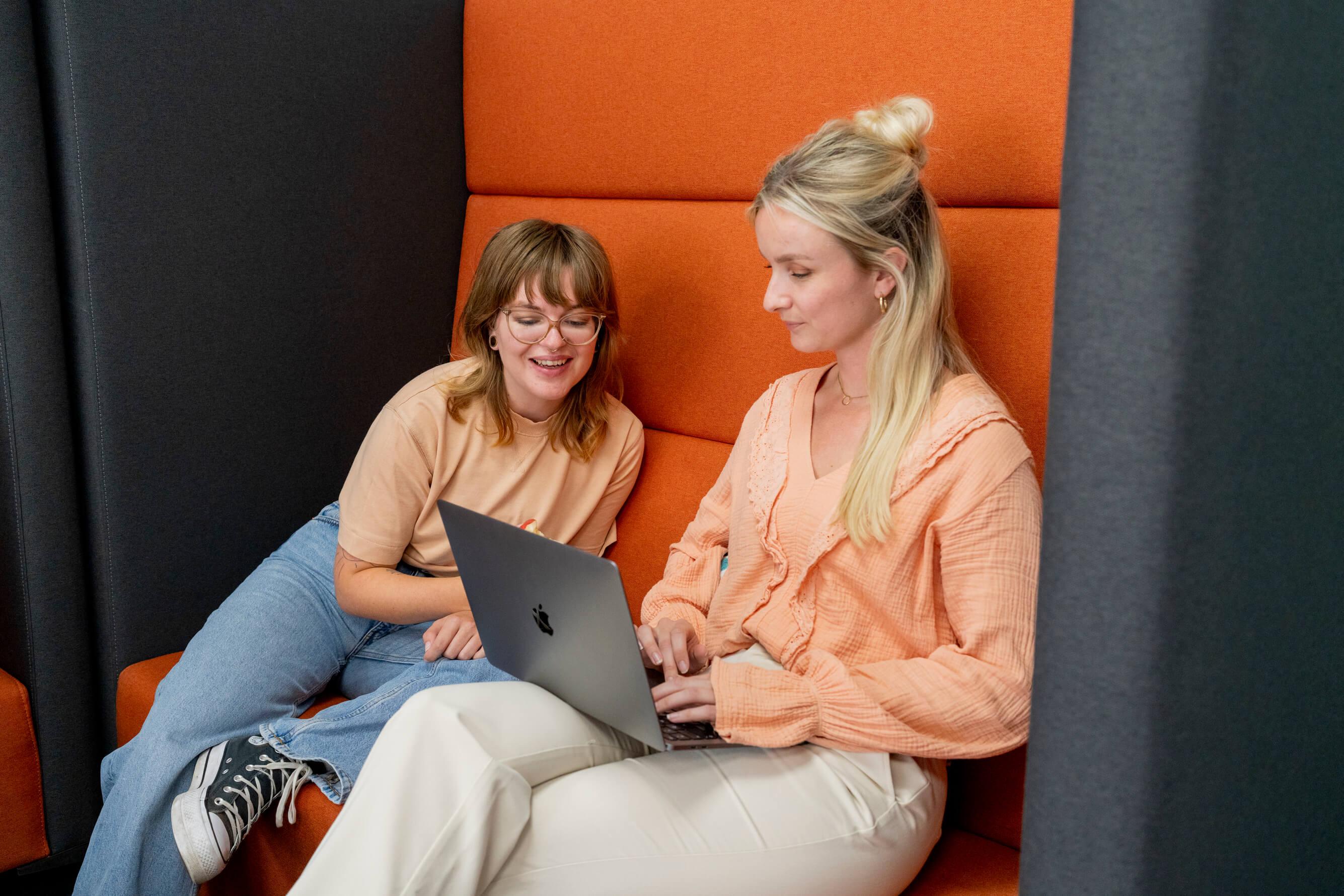 Twee vrouwen werken samen met een laptop in een oranje zithoek